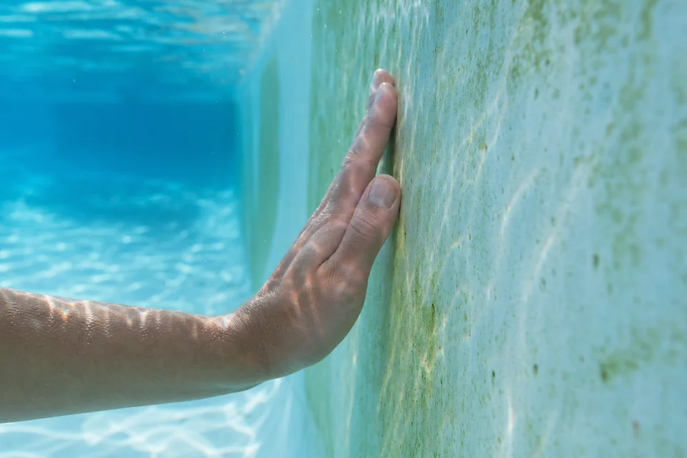 Hand prüft den glitschigen Biofilm an einer Poolwand unter Wasser