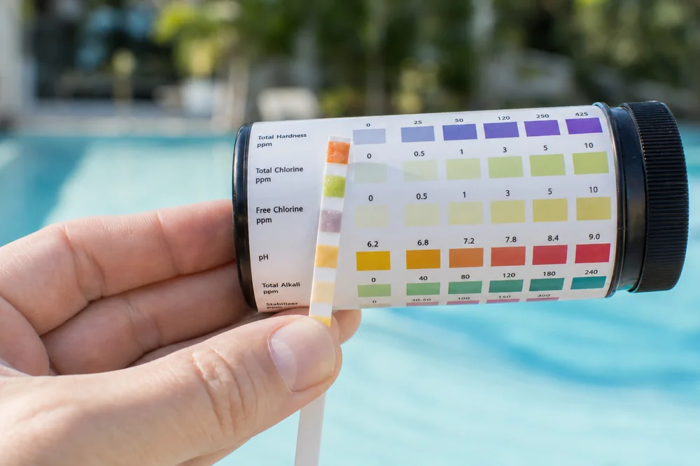 Hand hält Teststreifen vor eine Farbvergleichsskala am Pool