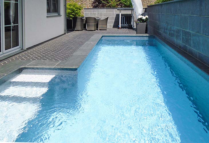 Draufsicht auf ein rechteckiges Betonschwimmbecken mit fertig verlegter Poolfolie, eingefasst von dunkler Granitumrandung, integrierten Stufen im Becken und Kies- sowie Gräserbepflanzung am Beckenrand.