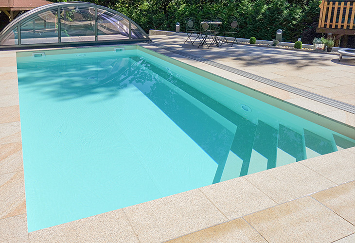 Hellblau ausgelegte Poolfolie in einem rechteckigen Betonpool mit integrierten Treppen und Sitzbank, eingefasst von hellen Travertinplatten und minimalistisch gestalteter Gartenumgebung.