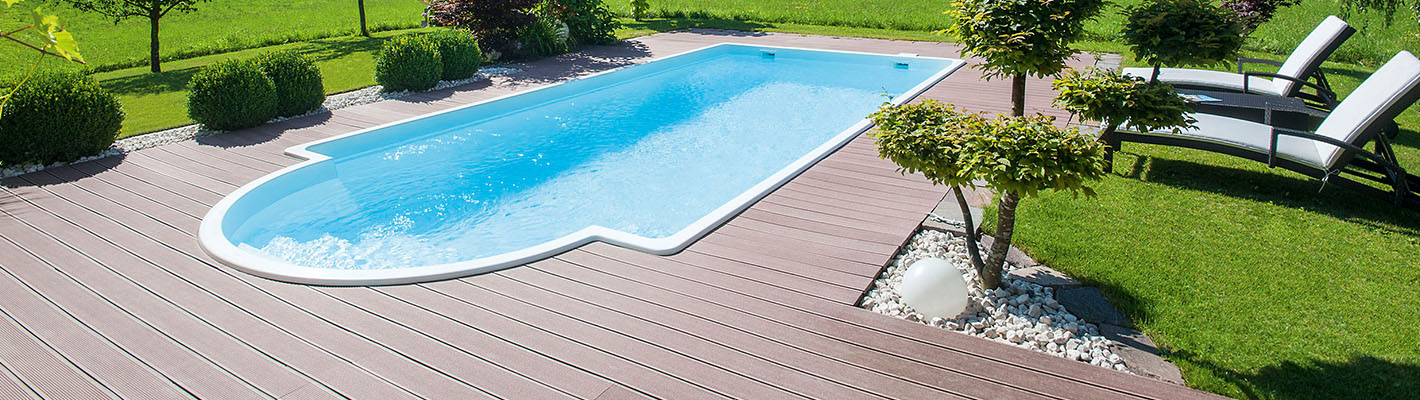 GFK Schwimmbecken Polyfaser – Eleganter Gartenpool mit Holzdeck GFK Schwimmbecken aus Polyfaser mit geschwungener Einstiegsstufe, umgeben von einer WPC-Holzterrasse, dekorativen Kiesflächen mit kleinen Bäumen, Rasen und zwei Relax-Liegen im Hintergrund.