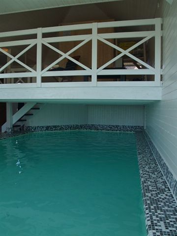 Hallenbecken mit Mosaikfliesen am Beckenrand und darüberliegendem Balkon mit weißem Geländer