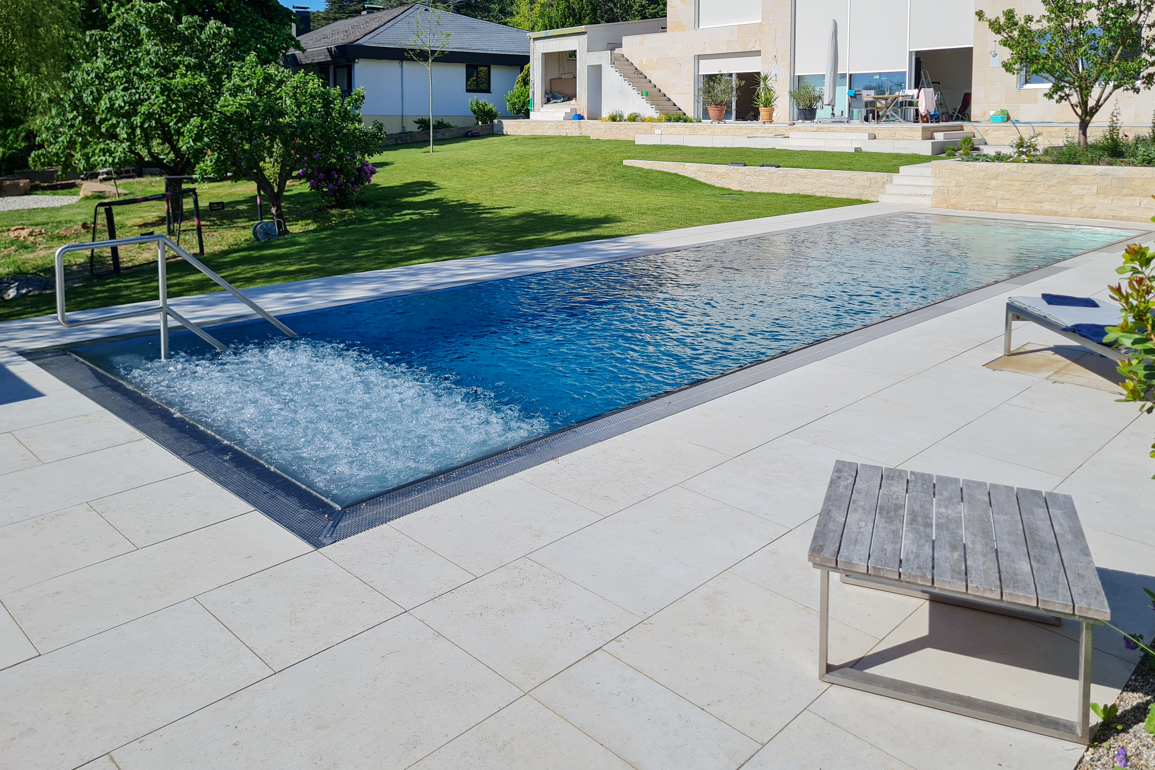 Langgezogener Edelstahlpool mit Luftsprudelliege auf voller Breite und Blick auf den Garten mit moderner Außengestaltung