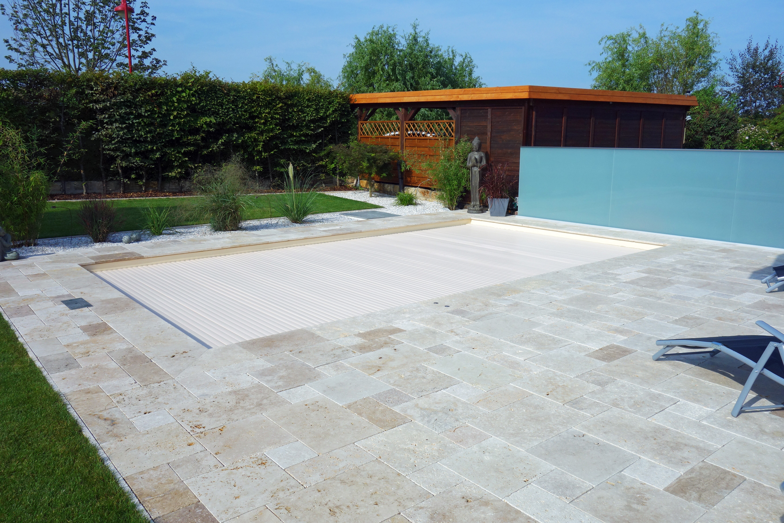 Außen-Pool mit geschlossener Rollladenabdeckung, umgeben von einer hellen Steinterrasse und grüner Bepflanzung.