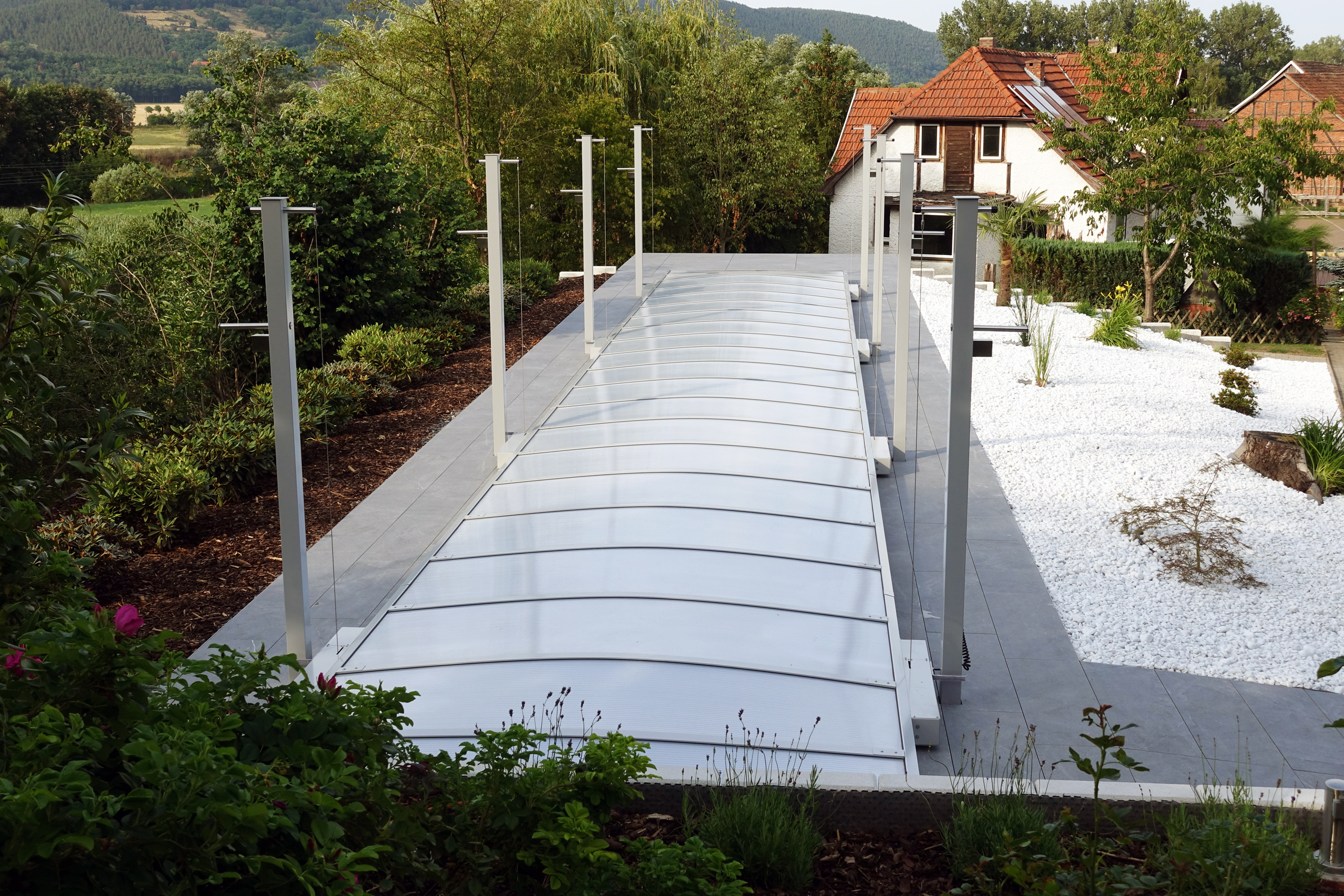 Schwimmbecken mit geschlossenem Hubdach in einer Gartenanlage, umgeben von Bäumen und einem Haus im Hintergrund.