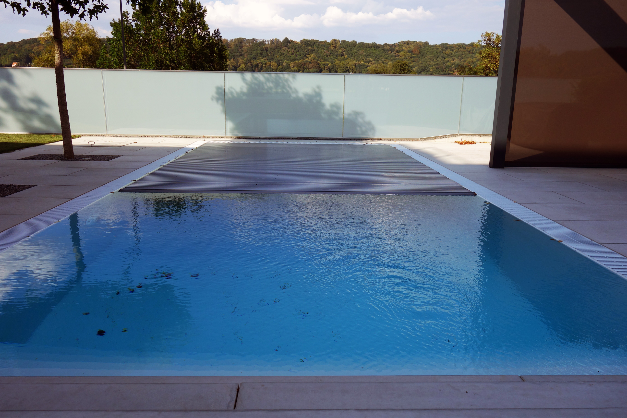 Überlaufpool mit blauer Folie, teilweise abgedeckt von einem Unterflurrollladen, umgeben von hellen Fliesen.