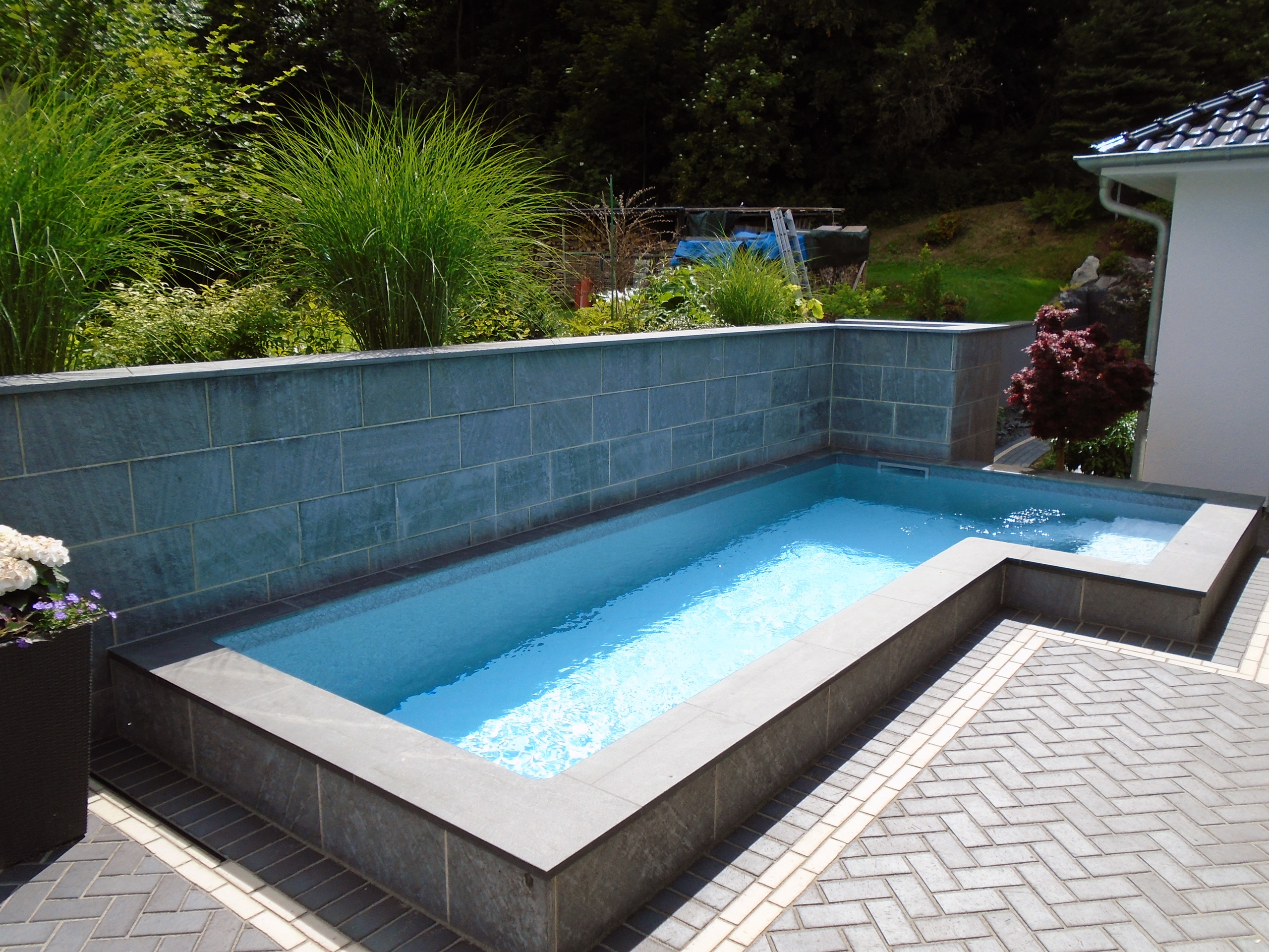 L-förmiges Schwimmbecken mit Treppe und Podest, umgeben von einer gepflasterten Terrasse und Bepflanzung.