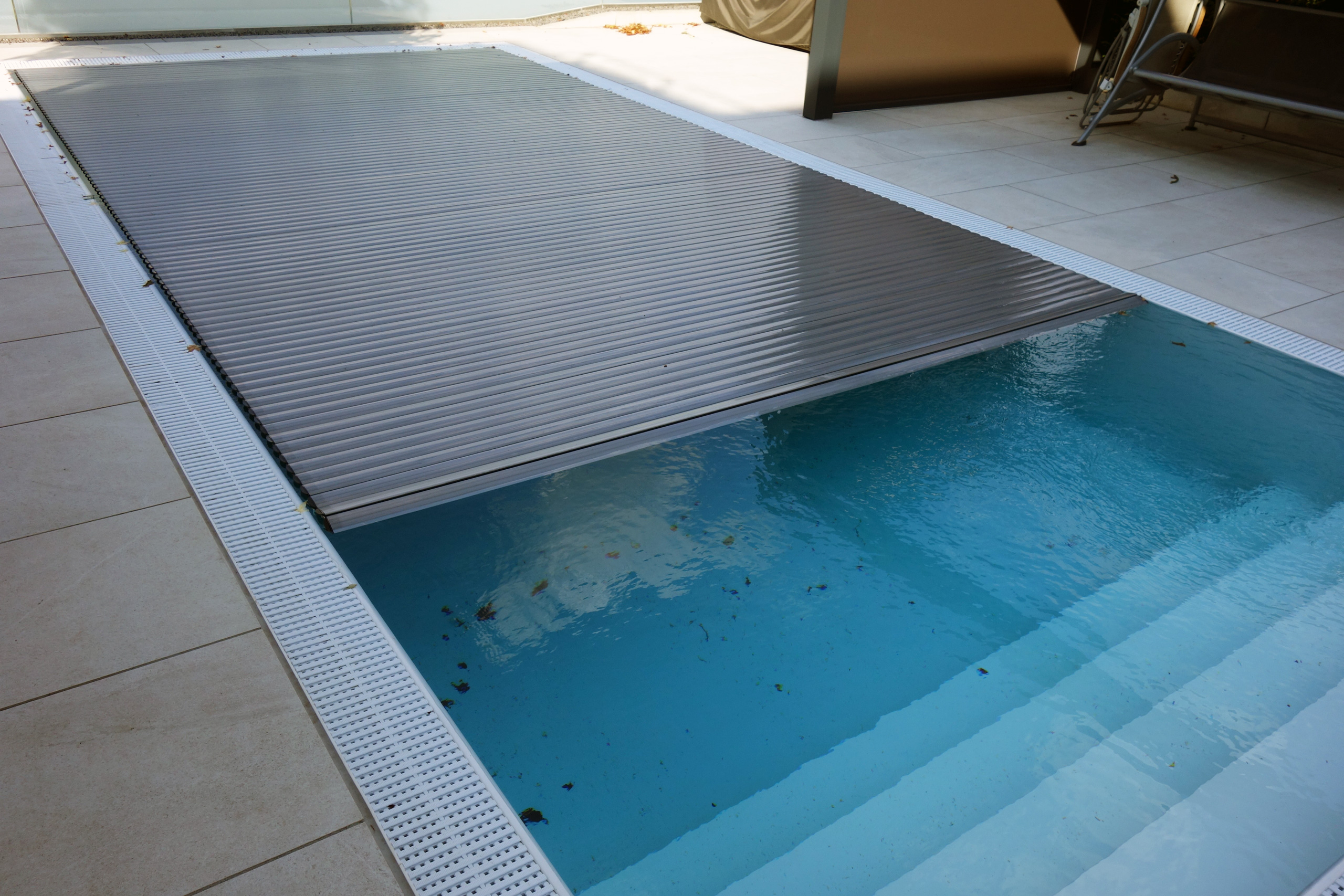 Teilansicht des Überlaufpools mit Unterflurrollladen und der geraden Treppe, umgeben von hellen Fliesen