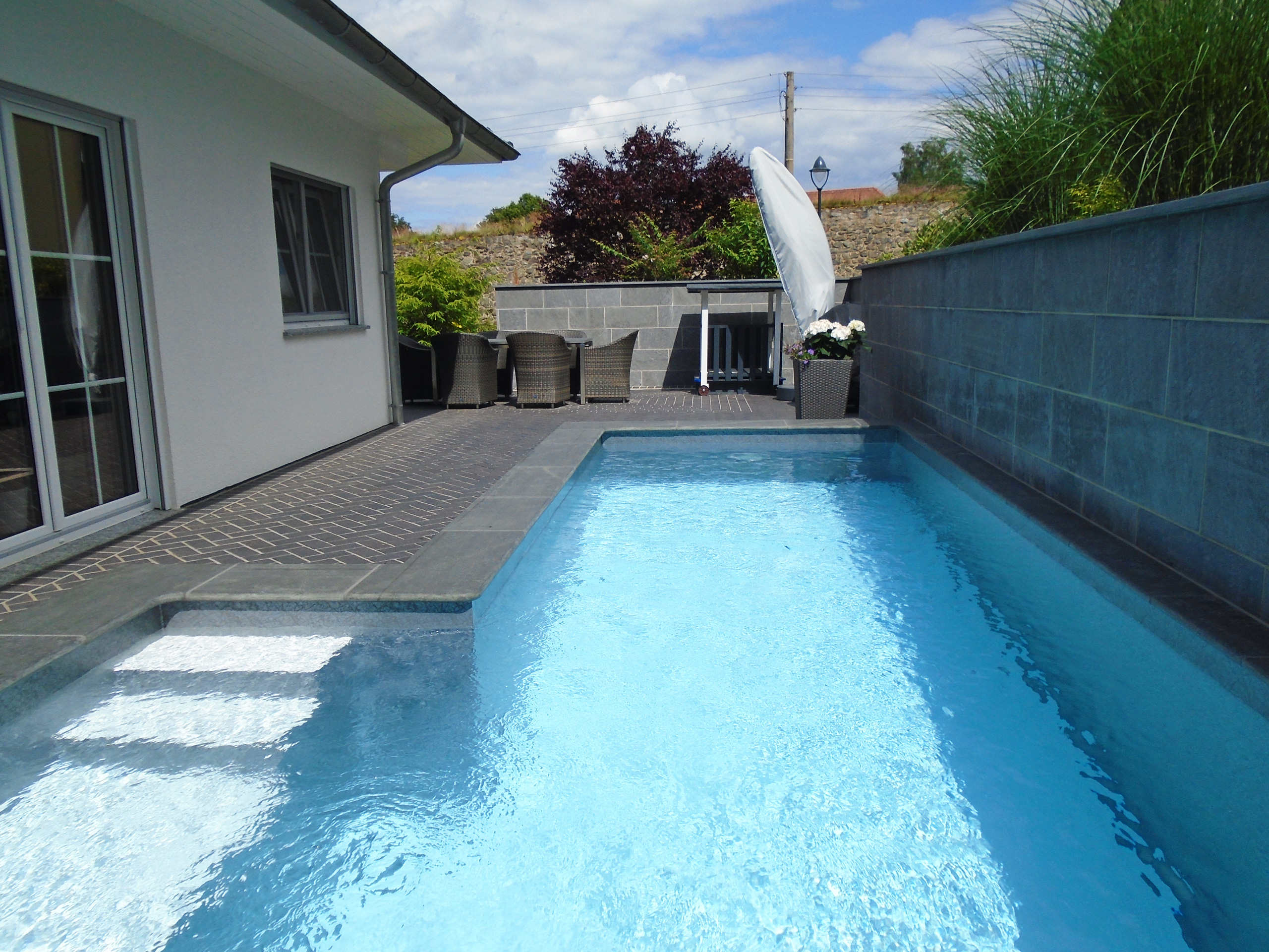 L-förmiges Schwimmbecken mit Treppe und Podest, umgeben von einer Terrasse und Hauswand.