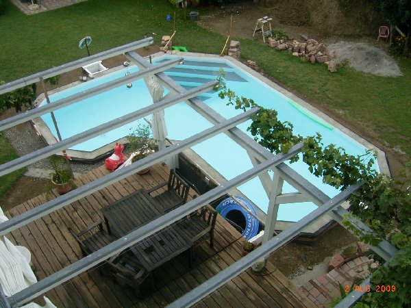 L-förmiges Schwimmbecken mit trapezförmiger Treppe, von oben betrachtet, umgeben von Terrasse und Garten