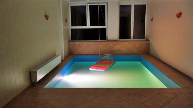 Hallenbecken mit blauer Einhängefolie und Unterwasserbeleuchtung, ein Schwimmbrett schwimmt auf dem Wasser.
