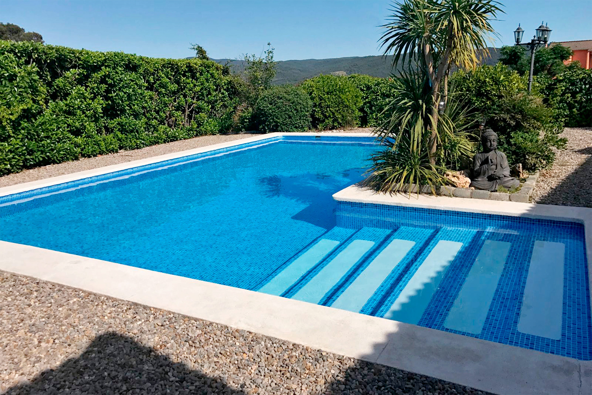 Schwimmbad mit ELBE Supra Print Schwimmbadfolie in Mosaik Blau und integrierter Treppe, umgeben von Kies und Pflanzen.