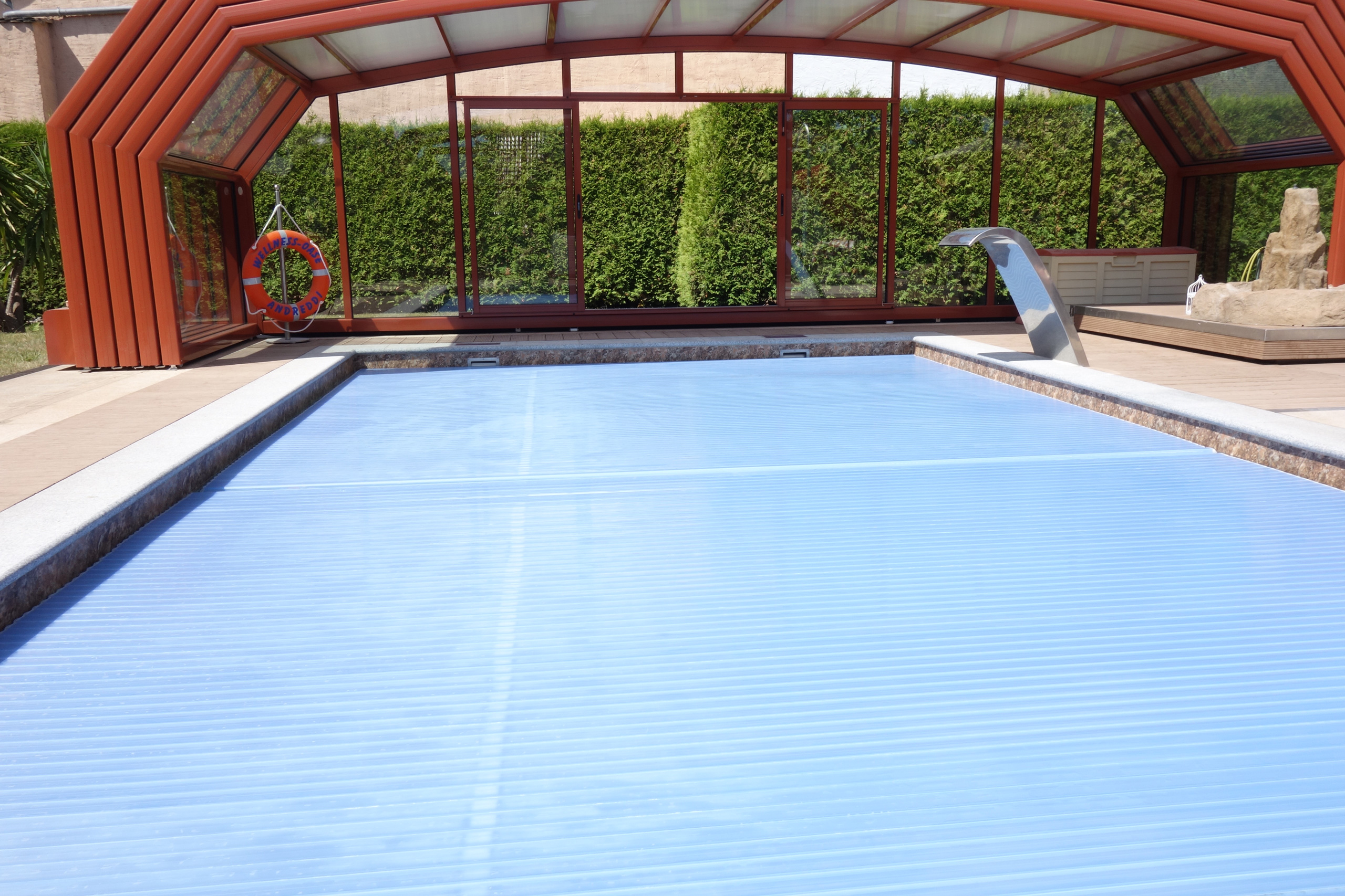Schwimmbecken mit blauer Rollladenabdeckung und braun-roter Poolüberdachung, im Hintergrund eine Hecke.