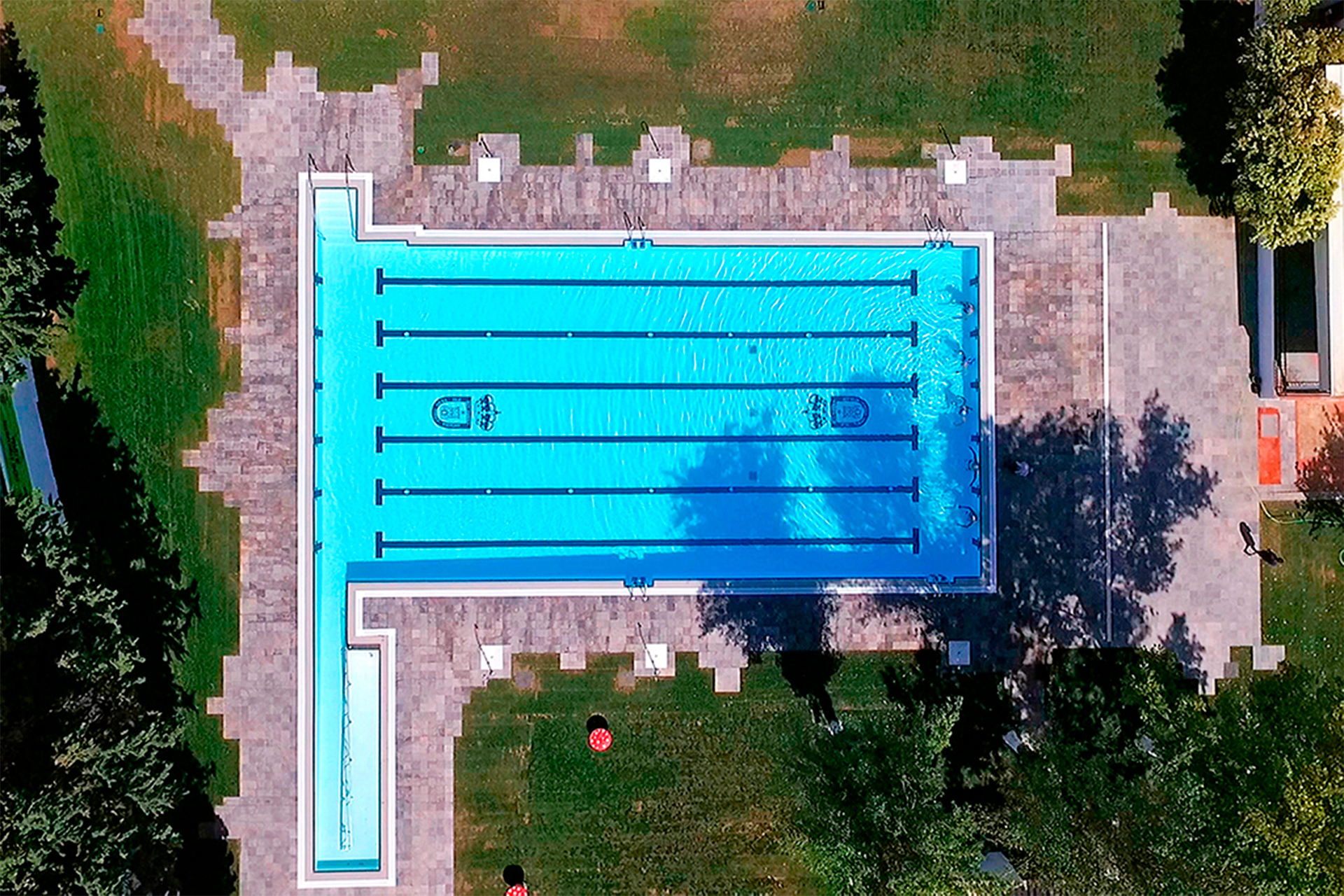Luftbild einer großen Schwimmbadanlage mit Bahnen und angrenzendem Becken, umgeben von Grünflächen und Wegen.