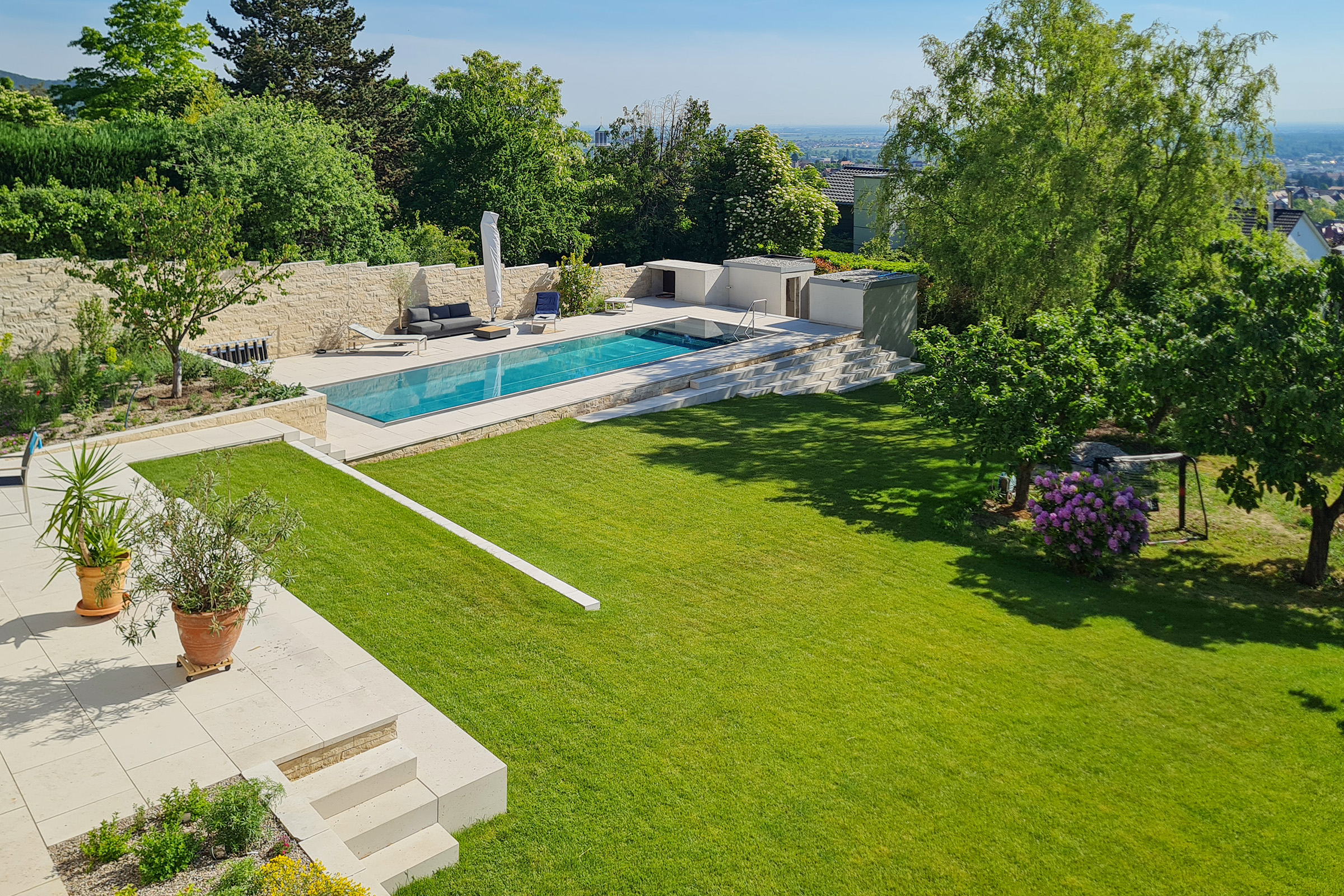 Länglicher Edelstahlpool mit gerader Treppe und Blick über die Stadt, umgeben von Grünflächen und Terrasse