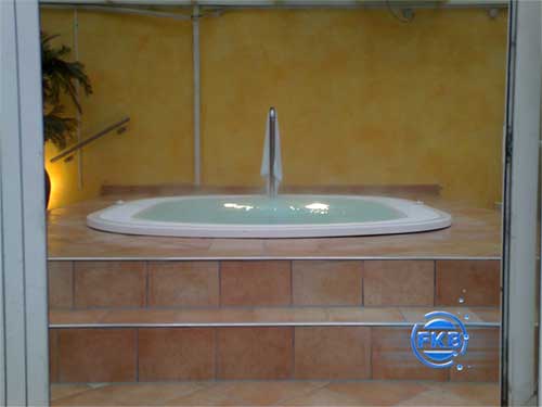 Eingelassener Whirlpool MANDURA mit Wasserspeier, umgeben von braunen Fliesen und gelber Wand.